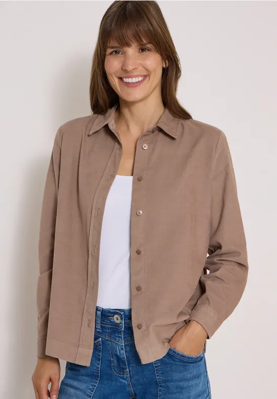 Une femme souriante porte une veste marron sur un débardeur blanc et un jean.