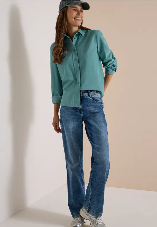 Femme souriante avec chemise verte, jean et casquette, adossée à un mur.