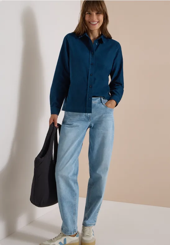 Femme en chemise bleu foncé, jean clair et baskets claires avec un sac noir, posant devant un mur neutre.