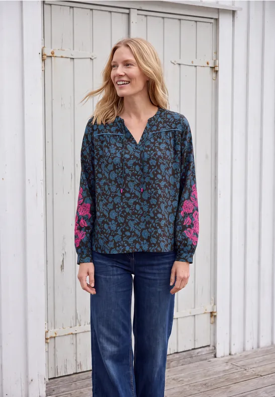 Eine lächelnde Frau in blau gemusterter Bluse mit pinken Ärmeln und Jeans vor einer weißen Holzwand.