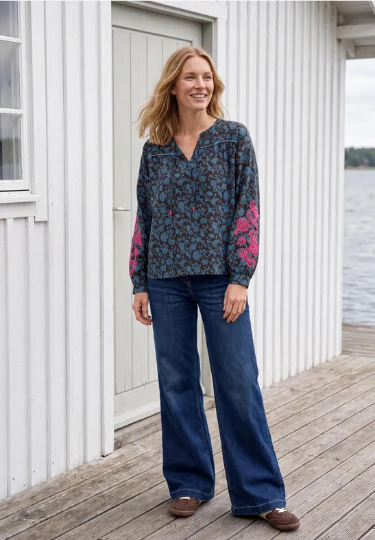 Eine lächelnde Frau in blauer, geblümter Bluse und Schlagjeans steht auf einem Steg vor einem weißen Haus am Wasser.