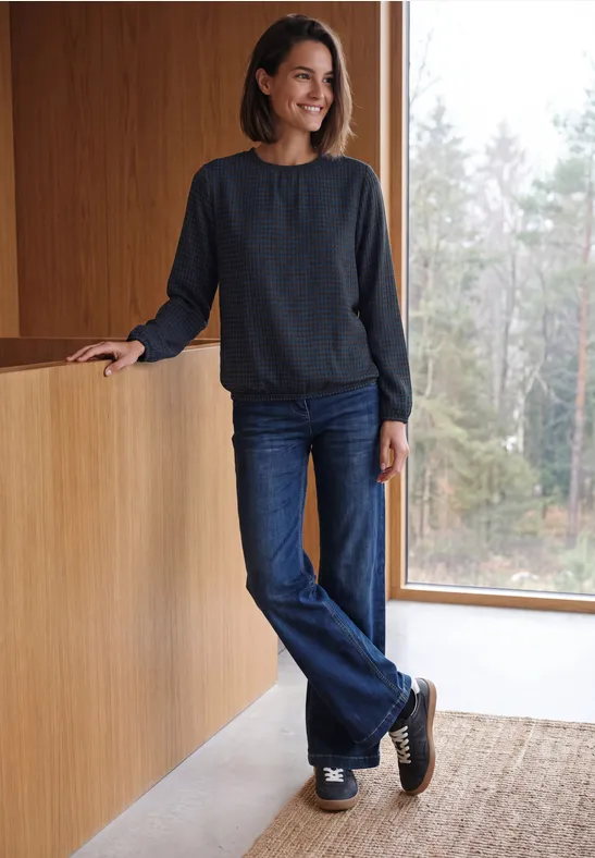 Eine lächelnde Frau in Jeans und dunklem Pullover lehnt an einer Holztheke und schaut aus dem Fenster.