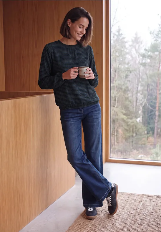 Eine Frau in grünem Pullover und Jeans steht an einem Fenster und hält eine Tasse in modernem Innenraum.