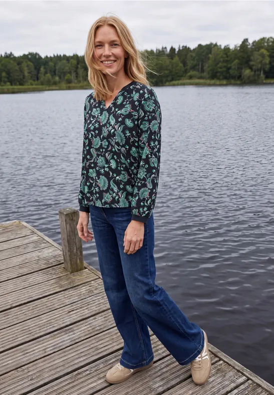 Une femme souriante sur un ponton au bord d’un lac, en chemisier à motifs et jeans.