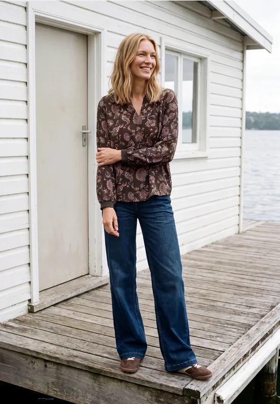 Lächelnde Frau in gemusterter Bluse und Jeans auf einem Steghaus am Wasser.