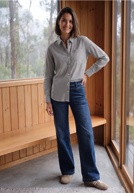 Femme dans une pièce en bois clair, souriante, portant une chemise grise, un jean bleu et des chaussures claires.