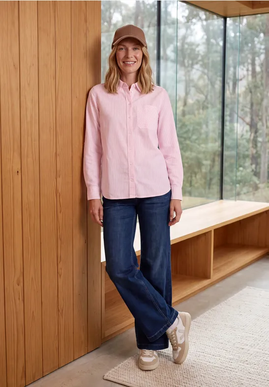 Frau in rosa Hemd, blauen Jeans und Mütze lehnt lächelnd an einer Holzverkleidung neben großen Fenstern.