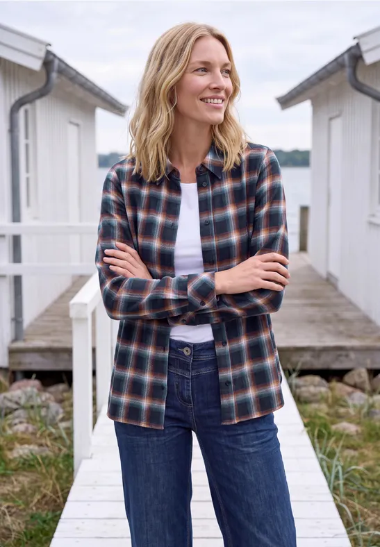 Une femme souriante en chemise à carreaux et jean se tient sur une passerelle au bord de l'eau.