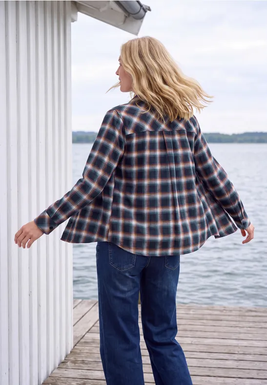 Femme en chemise à carreaux et jean marche sur une jetée en bois devant une cabane blanche au bord de l'eau.