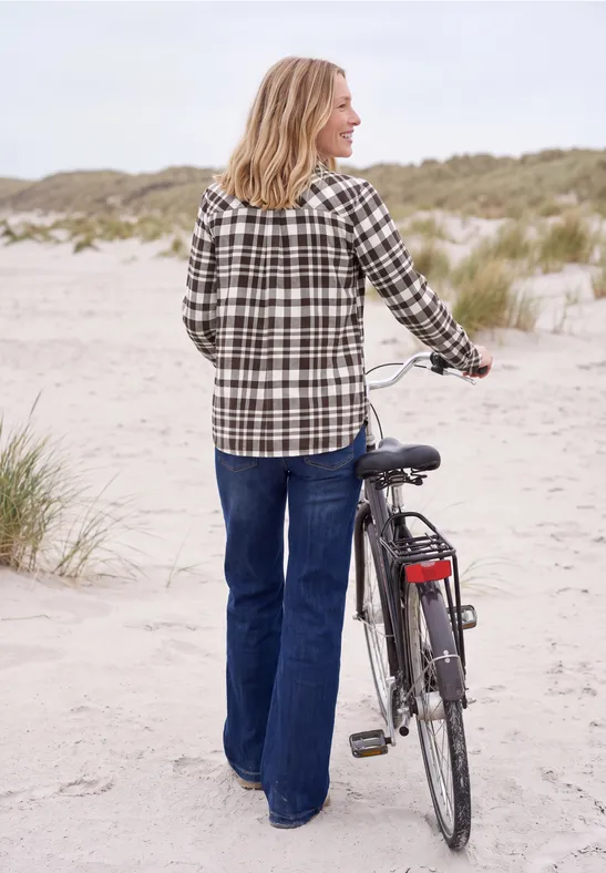 Frau in kariertem Hemd und Jeans schiebt Fahrrad am Dünenstrand entlang.