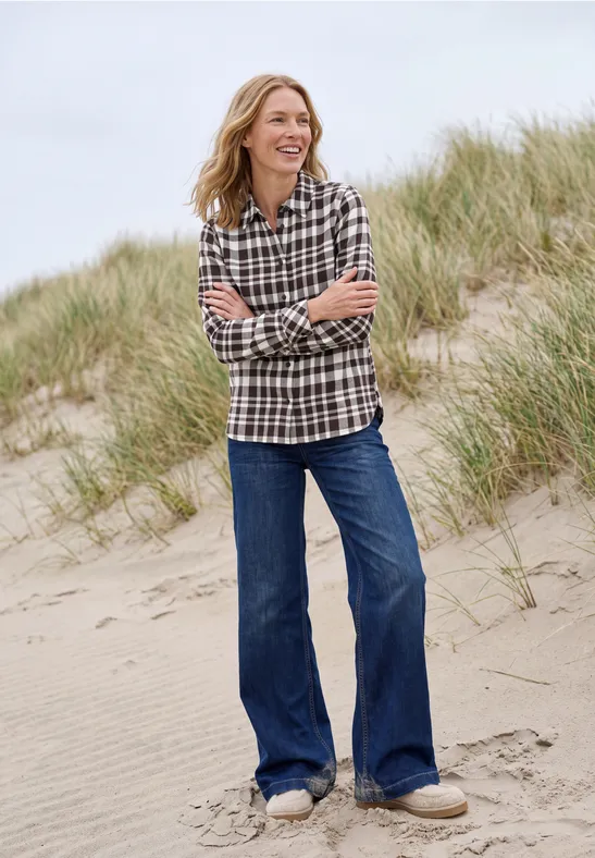 Eine lächelnde Frau in kariertem Hemd und weitem Jeans steht auf einer Sanddüne mit Strandgras.
