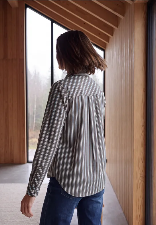 Eine Frau trägt ein gestreiftes Hemd und Jeans, steht seitlich in einem Holzraum und blickt aus dem Fenster.