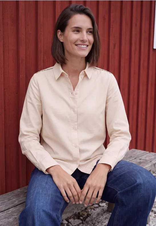 Une femme souriante assise sur un banc en bois devant un mur rouge, vêtue d'une chemise beige et d'un jean bleu.