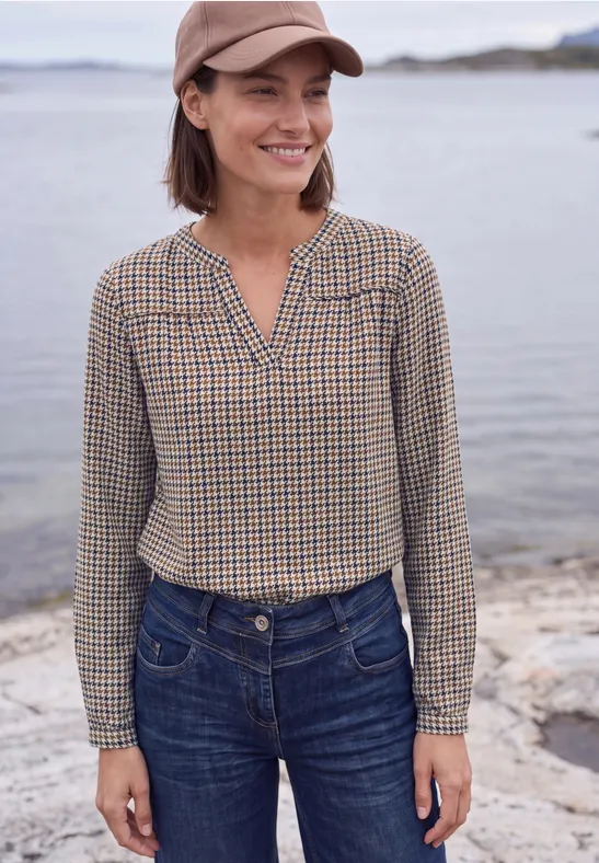 Frau mit kariertem Hemd, Jeans und Baseballkappe steht lächelnd am Ufer.
