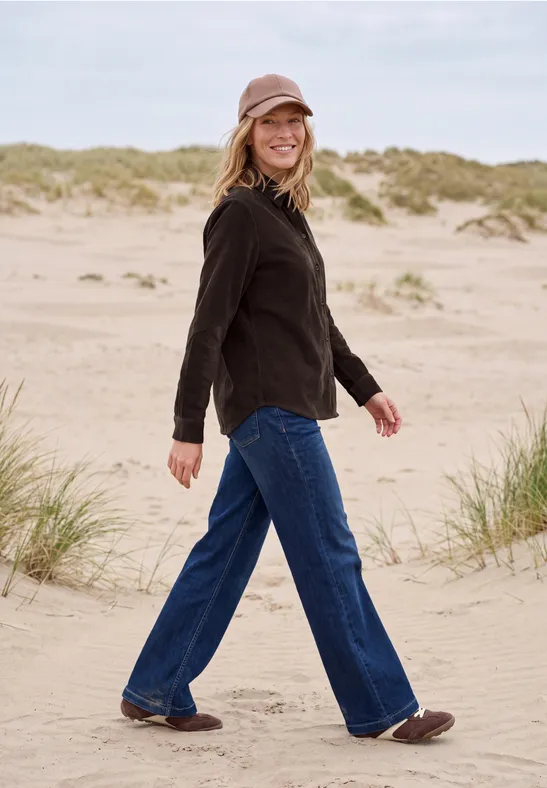 Frau in schwarzer Jacke, Jeans und Mütze spaziert lächelnd über Sanddünen am Strand.