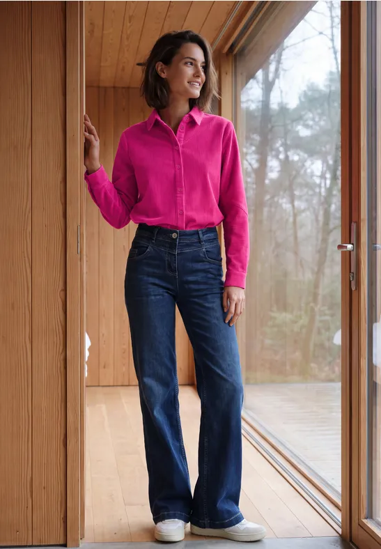 Frau in pinker Bluse und blauen Schlagjeans steht lächelnd in einem hellen Holzraum am Fenster.