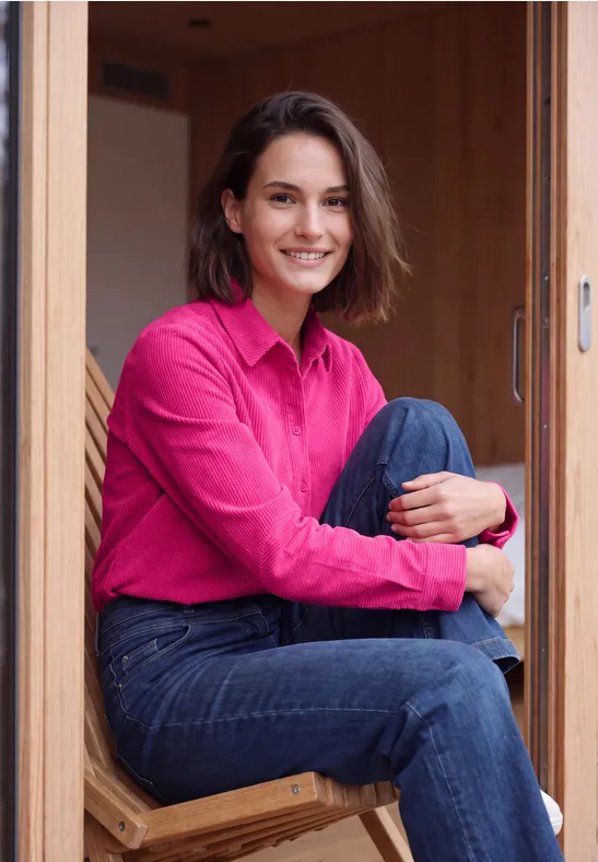 Eine Frau mit pinkem Hemd und Jeans sitzt im Holzrahmen und lächelt.