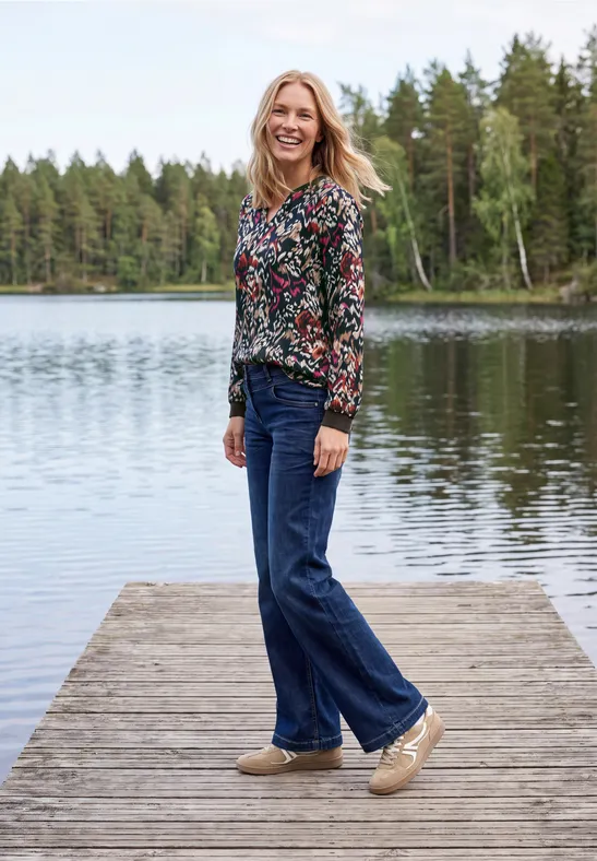 Une femme souriante est debout sur une jetée en bois au bord d’un lac paisible, forêt en arrière-plan.