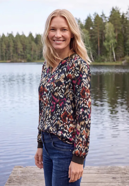 Femme souriante en chemisier coloré et jean, debout sur un ponton devant un lac et une forêt.