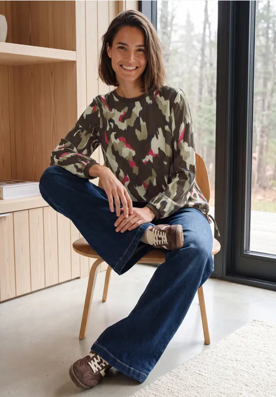 Eine Frau sitzt lächelnd auf einem Stuhl in einem hellen Raum, trägt Camouflage-Pullover und Jeans.
