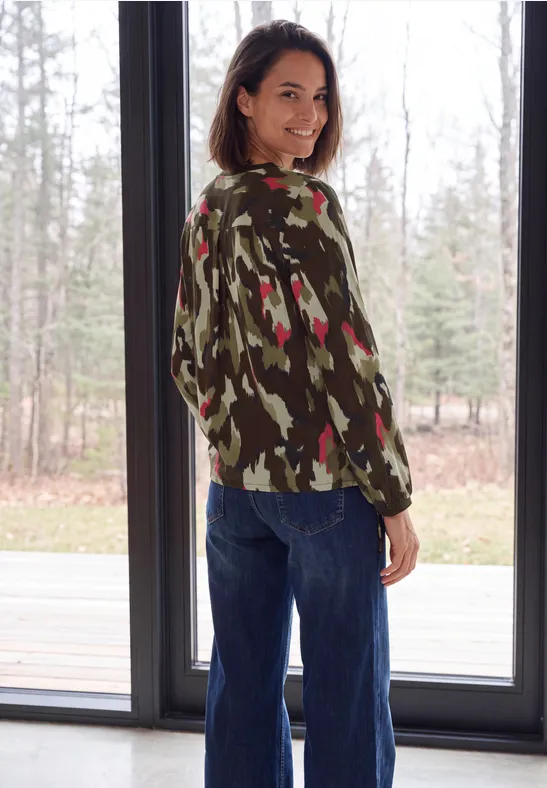 Eine lächelnde Frau im Camouflage-Pullover und Jeans steht vor einem großen Fenster und blickt über die Schulter.