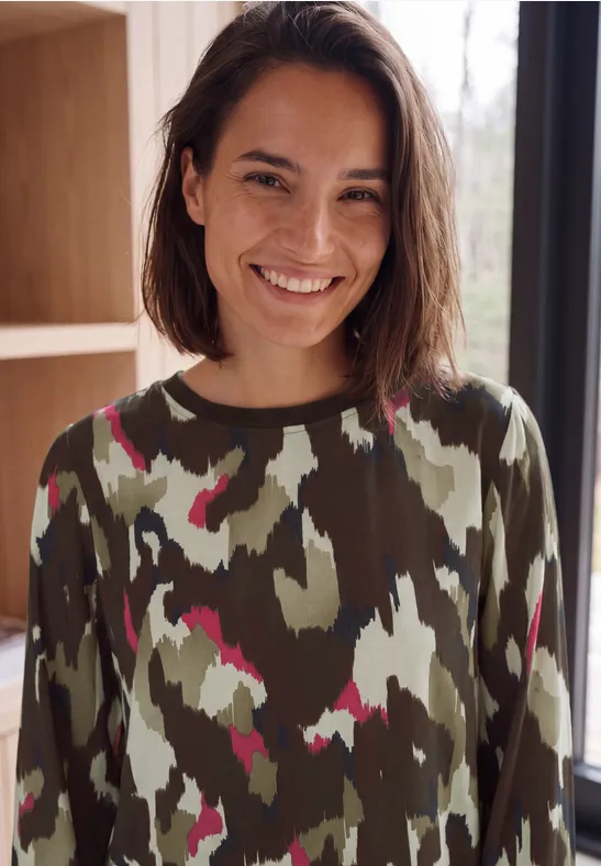 Junge Frau mit kurzem braunem Haar, lächelt in Camouflage-Pullover vor hellem Fenster.