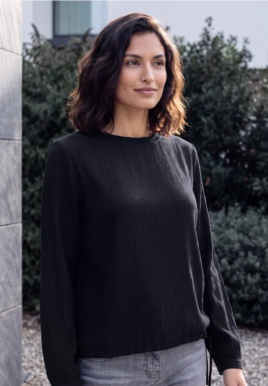 Eine lächelnde Frau mit schulterlangem dunklem Haar trägt eine schwarze Bluse und Jeans, steht vor Hecken und einer Steinwand.