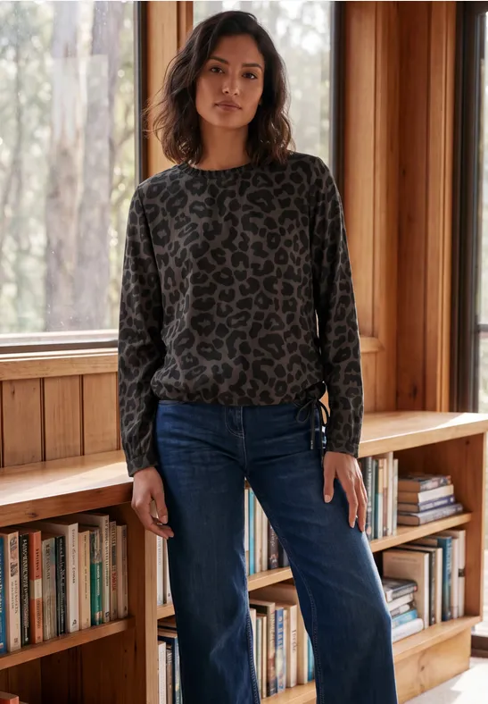 Frau in Leo-Pullover und Jeans steht vor einem Holzregal mit Büchern am Fenster.