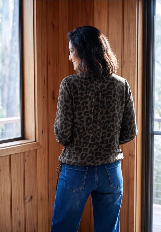 Eine Frau im Leopardenmuster-Oberteil und Jeans steht mit dem Rücken zur Kamera und blickt aus einem Holzfenster.