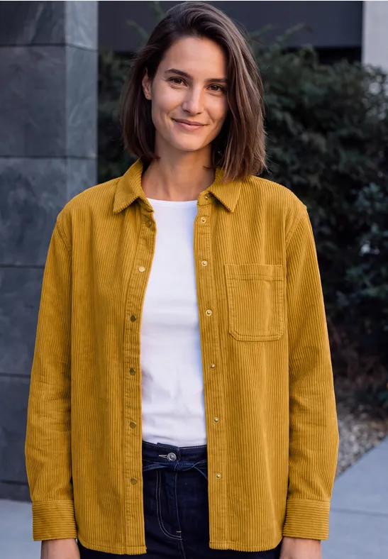 Une femme souriante se tient devant un bâtiment, portant une chemise moutarde, un t-shirt blanc et un jean foncé.