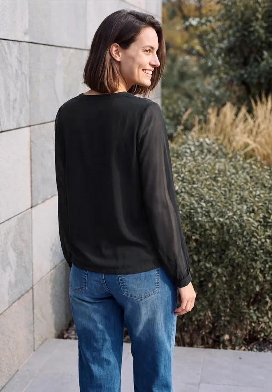 Frau in schwarzer Bluse und Jeans, steht lächelnd seitlich an einer Mauer, im Hintergrund Pflanzen.