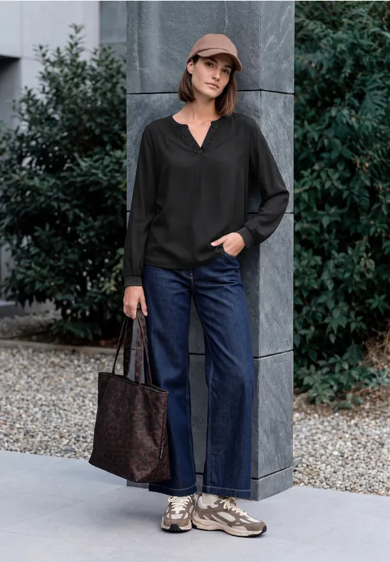 Frau mit Cap, schwarzer Bluse, Jeans und Sneakers posiert mit Tasche vor moderner Hausfassade.