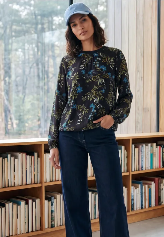 Junge Frau in lässigem Outfit mit gemustertem Pullover, weiter Jeans und Cap vor einem Bücherregal und großem Fenster.