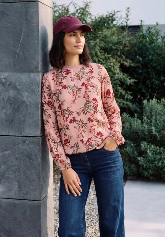 Eine Frau lehnt draußen an einer Säule, trägt eine rosa Blumenbluse, dunkelblaue Jeans und eine bordeauxrote Mütze.
