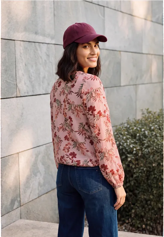 Frau in Blumenbluse und Jeans, trägt eine rote Baseballkappe und lächelt vor einer Mauer mit Grün.