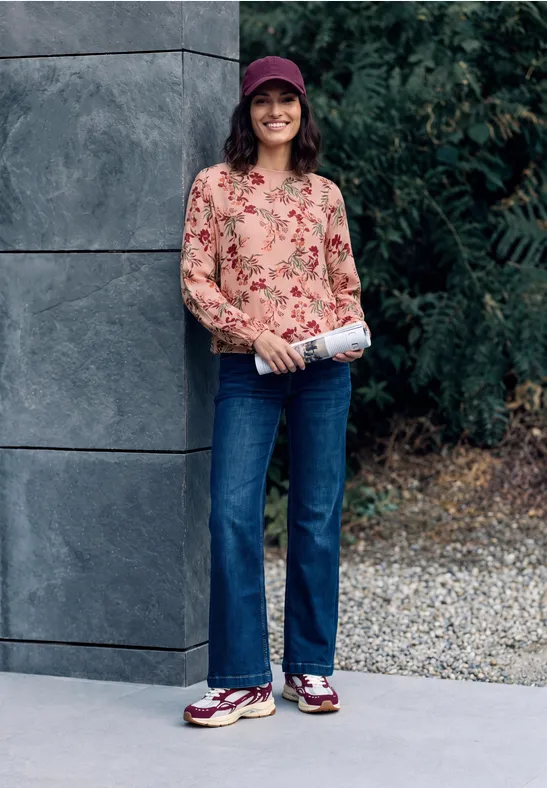 Eine Frau mit roter Cap, geblümter Bluse, Jeans und Sneakern lächelt und hält eine Zeitung.
