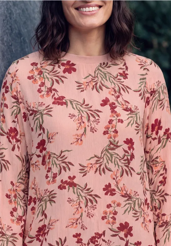 Frau trägt ein rosa Langarmshirt mit bunten Blumendrucken und lächelt sanft.