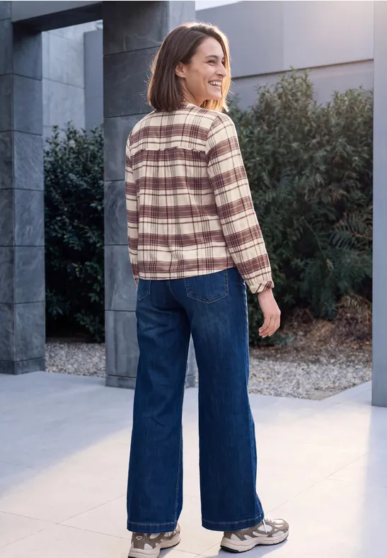 Femme devant un bâtiment moderne, portant une chemise à carreaux, jean large et baskets.