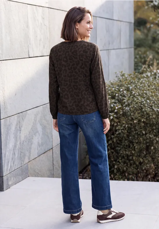 Une femme vue de dos porte un pull marron à motif, un jean bleu et des souliers bruns, adossée à un mur extérieur.
