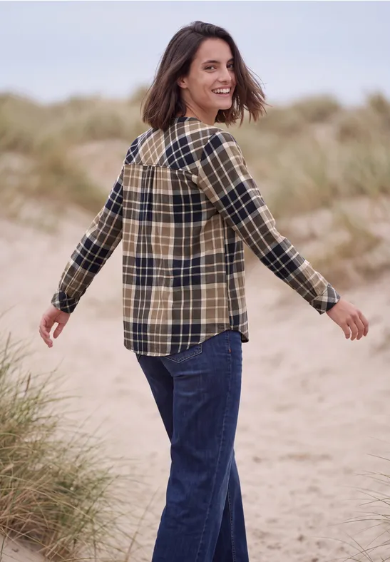 Eine lächelnde Frau in kariertem Hemd und Jeans spaziert am Strand und blickt über die Schulter.