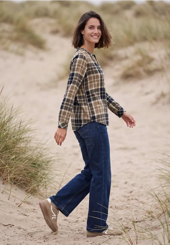 Eine Frau spaziert durch die Dünen, trägt karierte Bluse, blaue Jeans und flache Schuhe.