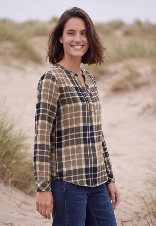 Eine Frau mit kariertem Hemd und Jeans steht lächelnd auf sandigen Dünen mit Gras im Hintergrund.