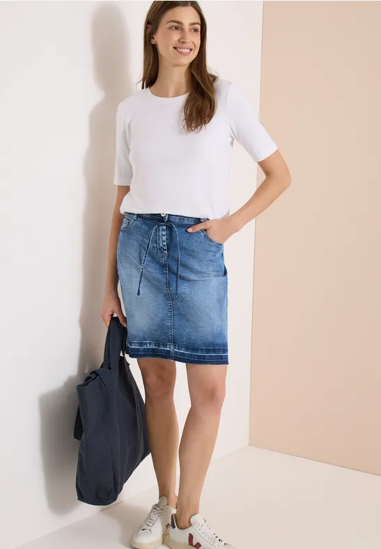 Frau vor heller Wand, trägt weißes T-Shirt, Jeansrock, weiße Sneaker, hält blaue Tasche.