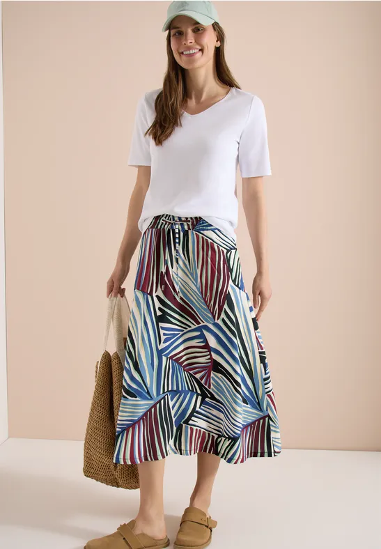 Frau trägt weißes T-Shirt, bunten gestreiften Midirock mit Volant, Strohtasche und Sandalen.