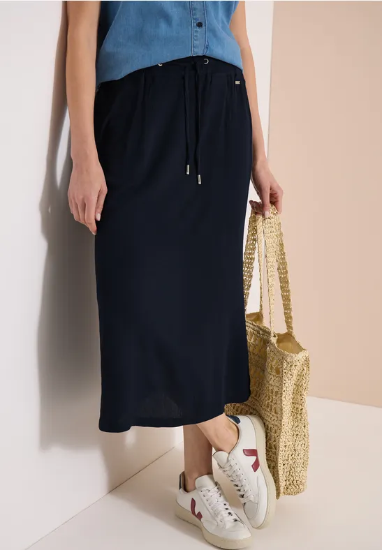 Frau trägt marineblauen Midi-Rock, weiße Sneaker und hält eine Strohtasche an der Wand.