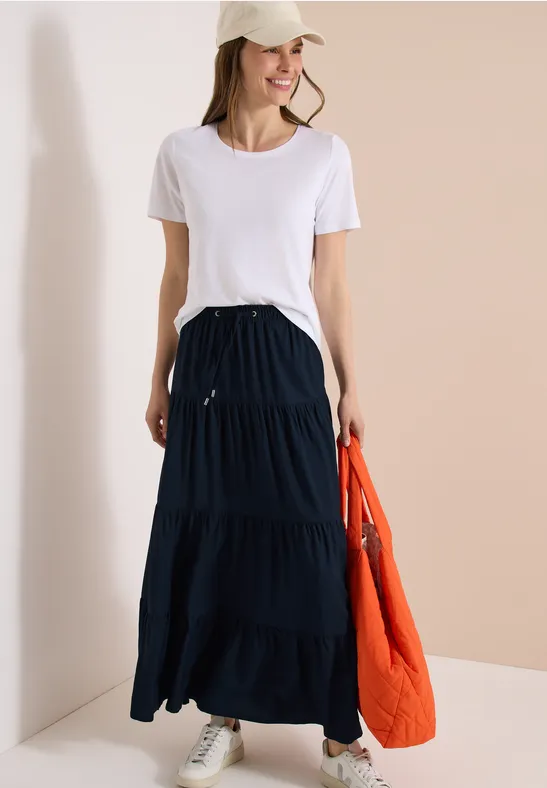 Femme en t-shirt blanc, jupe longue noire à volants, baskets grises, portant une doudoune orange.