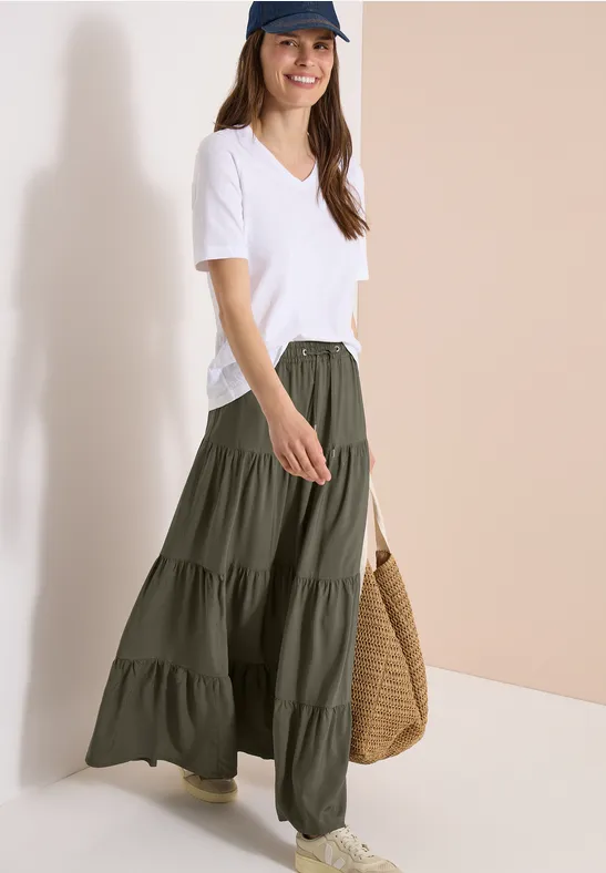 Frau in weißem V-Ausschnitt-Shirt und langem olivgrünem Stufenrock mit geflochtener Strohtasche.
