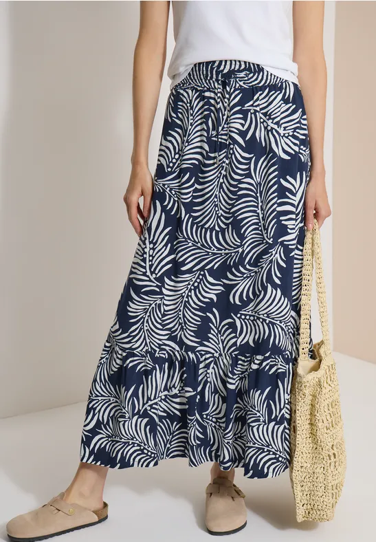Vrouw in wit topje en lange donkerblau met wit bladmotief, strooien tas en comfortabele schoenen.