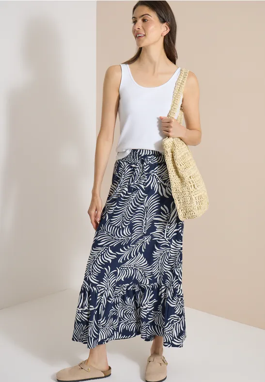 Vrouw in wit topje en lange blauw-witte rok met bladprint, rieten tas en sandalen voor twee tinten achtergrond.
