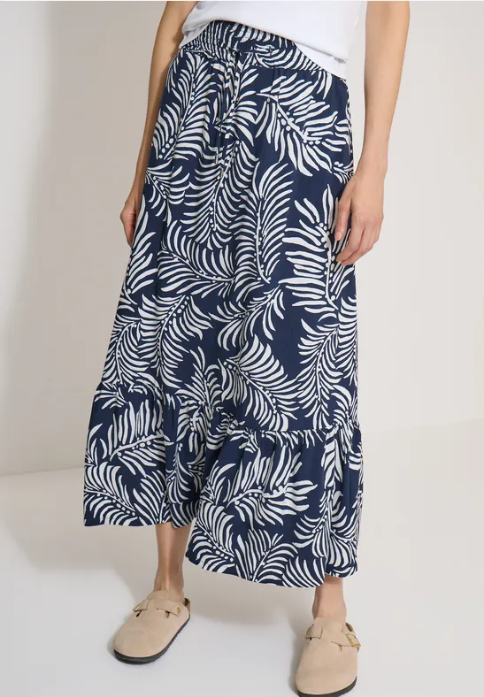 Vrouw draagt witte raffia instappers en een wijdvallende midirok met blauw-wit bladprint